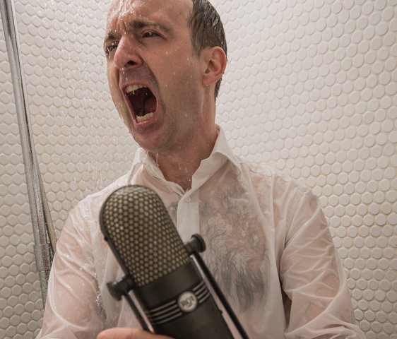 Patrick Bopp singt aus voller Kehle mit nasser Kleidung in einer Dusche, er hält ein Mikrofon in der Hand