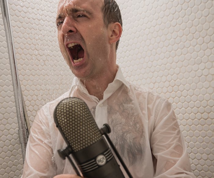 Patrick Bopp singt aus voller Kehle mit nasser Kleidung in einer Dusche, er hält ein Mikrofon in der Hand
